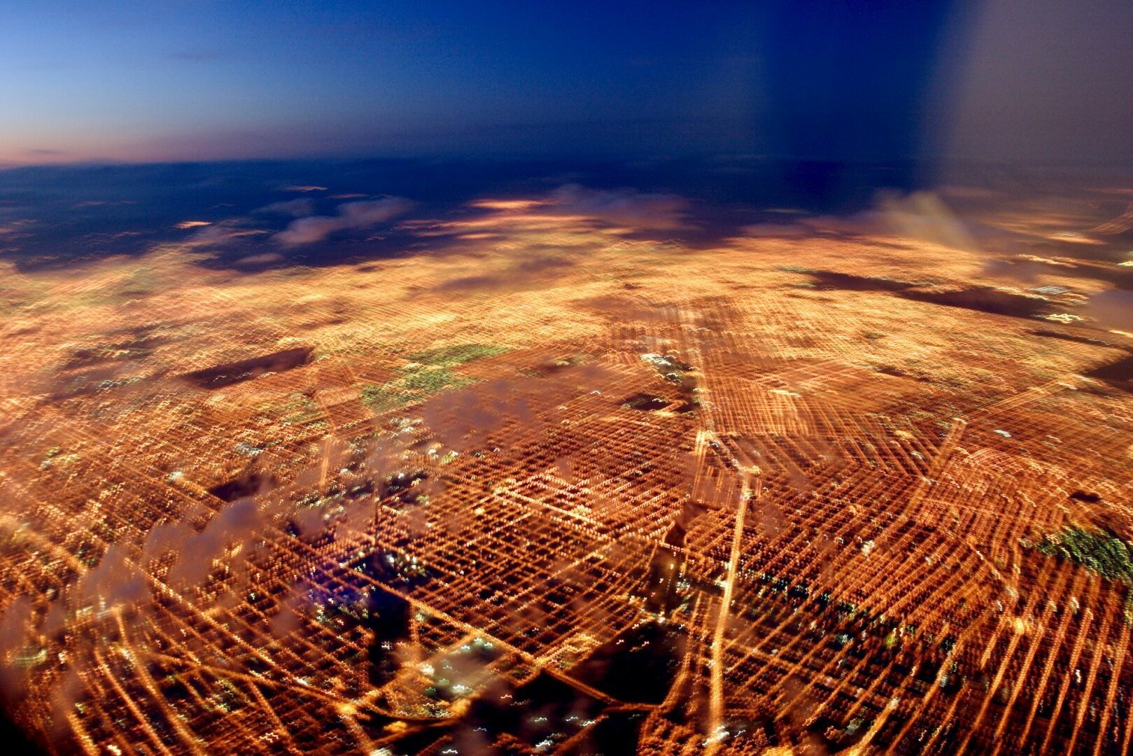 Aerial photo minutes before sunrise, there's some red color in the sky. Below is a huge city with roads and avenues grid lit in reddish/yellow light. The aircraft movement gives a subtle panning effect.