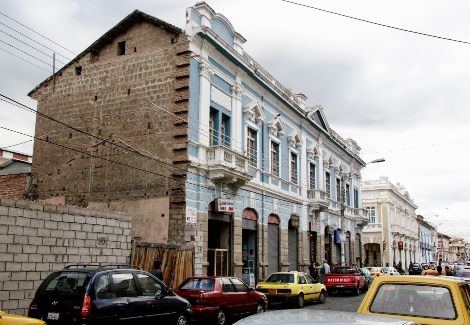 Image of a street in Riobamba, Ecuador. Overcast sky and dull light. XIX Century building with an ornate light blue and white facade but with a bare simple bricks lateral wall.