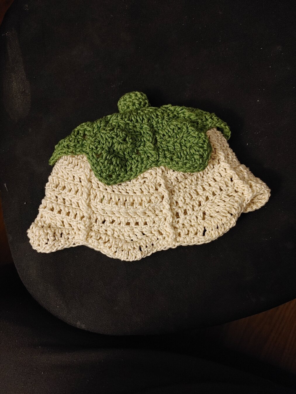 Crochet hat, toddler sized. It looks like a lily of the valley flower, with a green leafy top and white bottom with rounded edges. There is a small green stem on top.