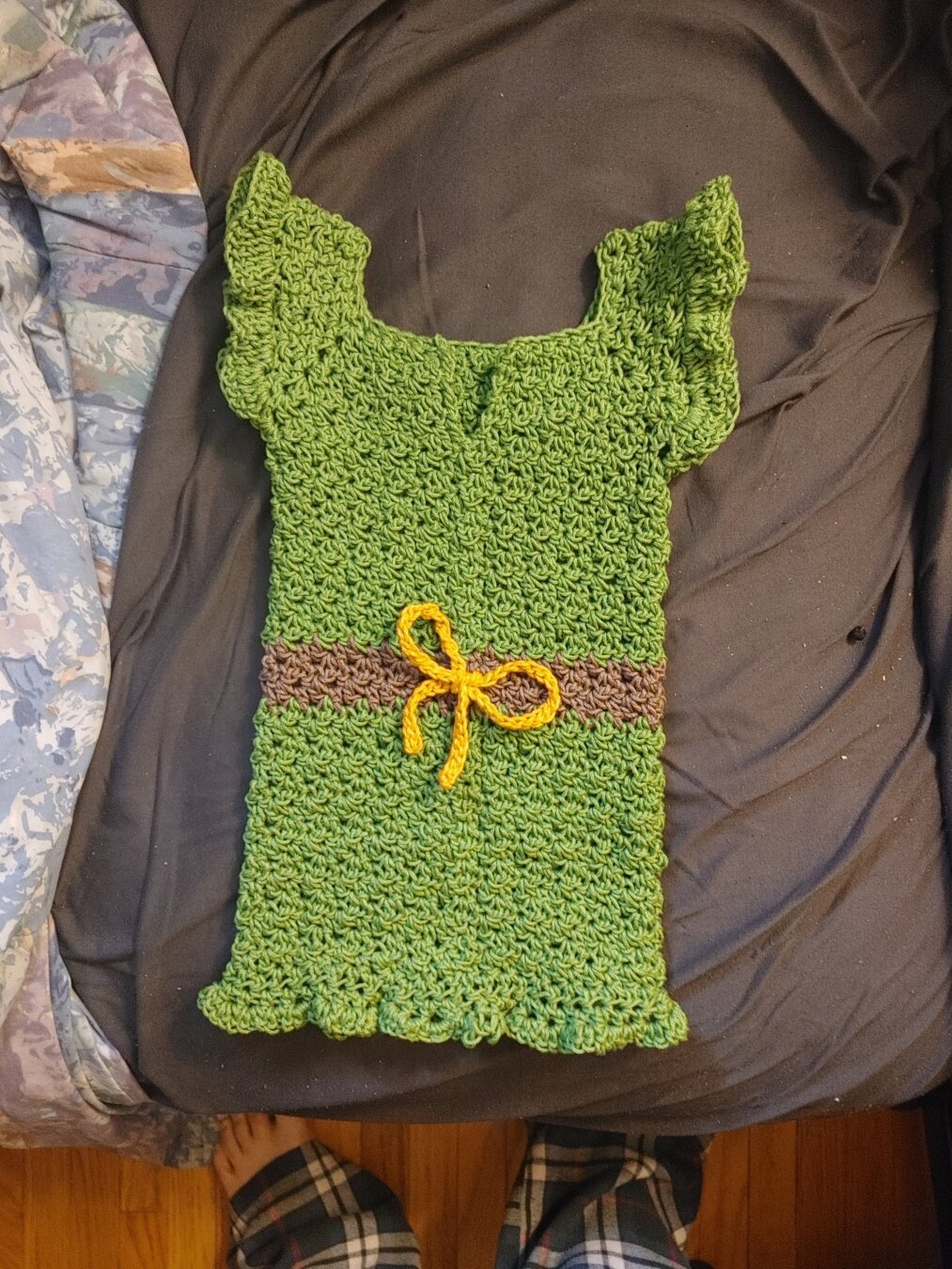Green crochet toddler dress with a brown horizontal stripe in the middle and a yellow bow, and a ruffled bottom and shoulders.