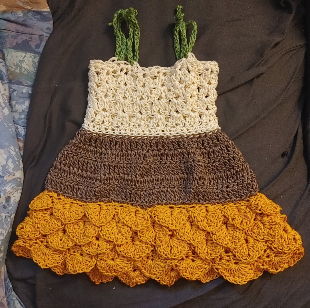 Crochet toddler dress with green tie on straps, beige top, brown middle, and a yellow skirt with crochet sunflower petals.