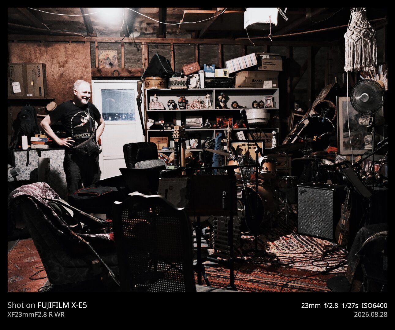 A musician standing in a densely cluttered garage practice space. There are guitars, chairs, a drum set, music stands, a framed poster leaning against a wall. It's chaos, but to the artist, it's delightful. (photo in color, with brown tones permeating the scene)