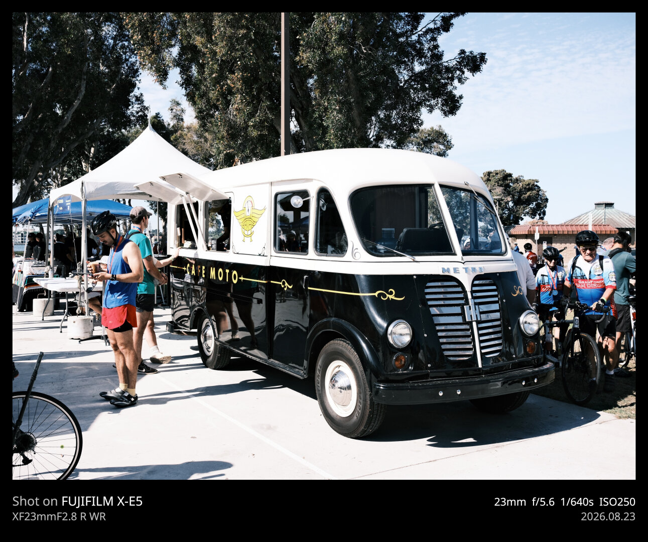 A classic Metro Coach bus with a black and cream color scheme has been converted into a mobile coffee shop. In the photo, a man is ordering from the window, while another man wearing a bike helmet looks at his phone. To the right, a couple of cyclists walk toward the frame.