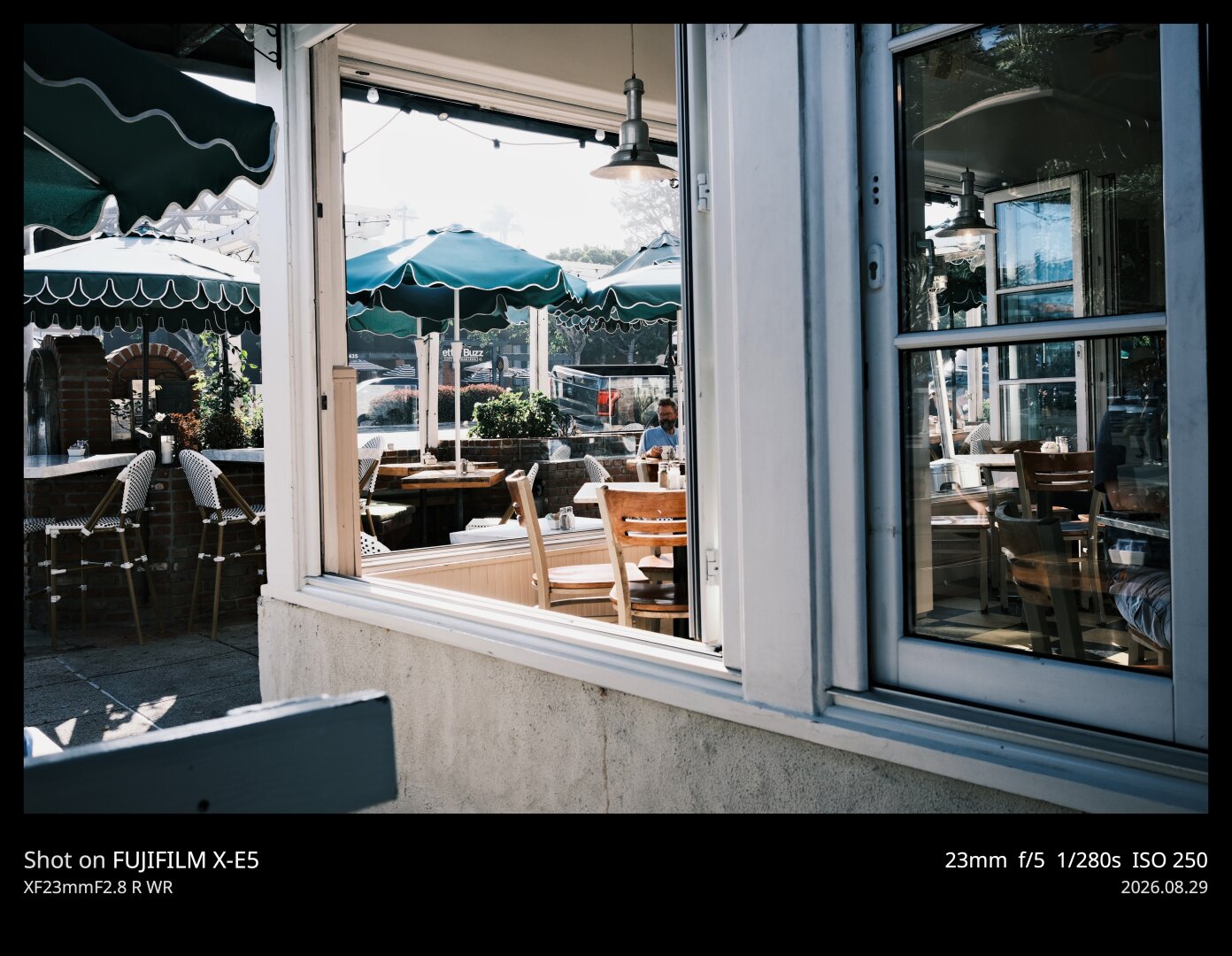 Point of view photo from a booth on the street outside of a diner in Del Mar, Southern California. Green umbrellas and accents all around, reflections in a window, bright morning sunlight and hard shadows.