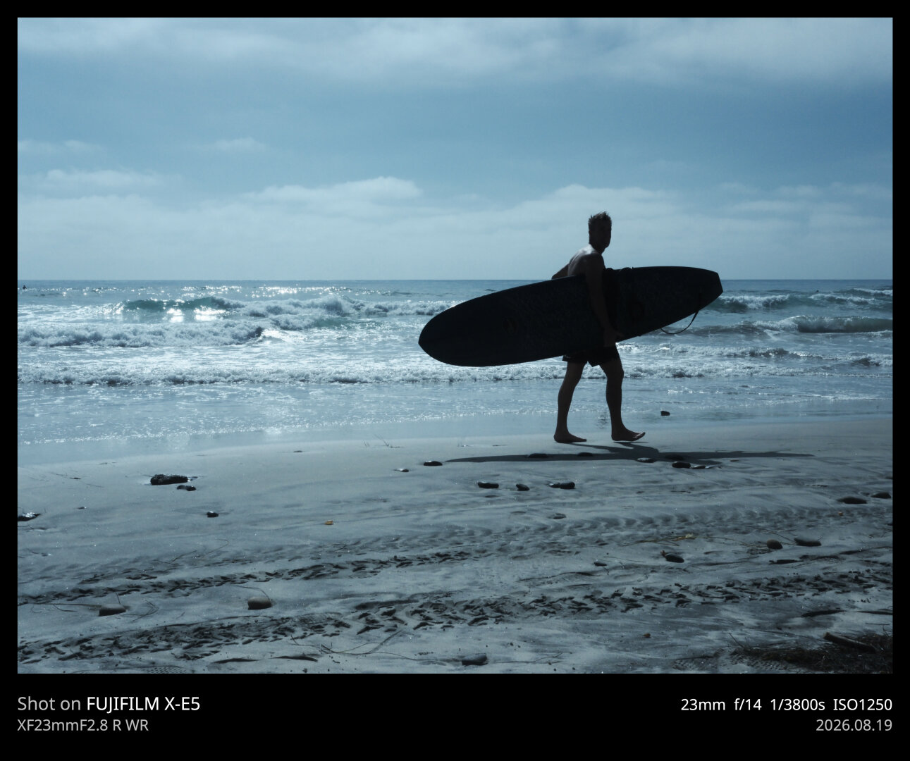 A surfer walks along the beach, silhouetted by the afternoon sun. The ocean is behind him, from the camera's point of view. The ocean and sky are starkly blue.