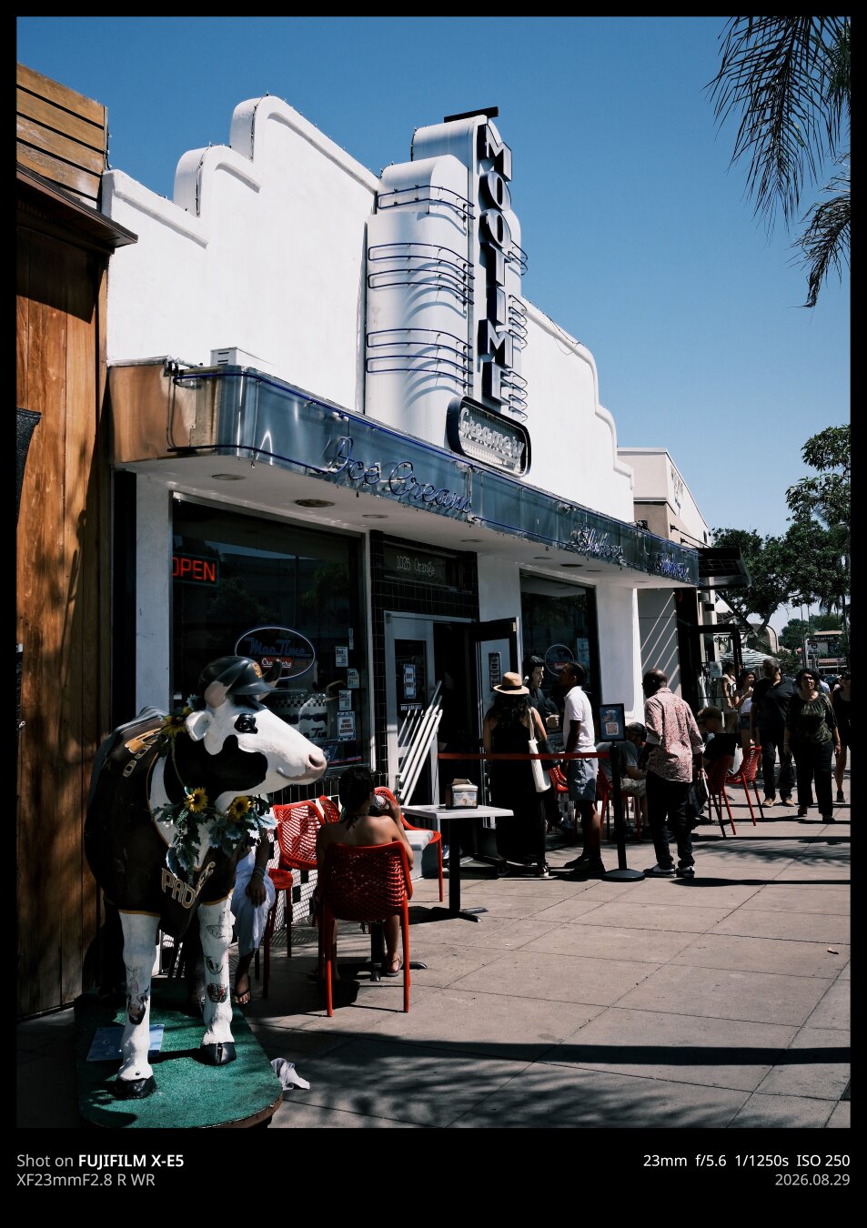 A photo of an art deco style ice cream shop in Coronado called "Mootime." The sign is a classic, vertical art deco affair made of metal. Out front is a cow statue wearing San Diego Padres gear. There's a line out the door of people wanting ice cream on a hot day.