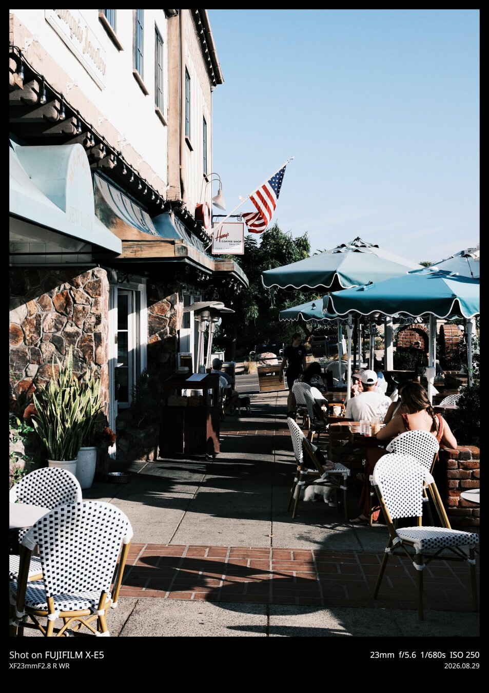 Portrait oriented photo showing the outside seating area of a diner in coastal southern California. There are green umbrellas, black and white checkered chairs, an American flag above, and hard shadows in the warm morning light.