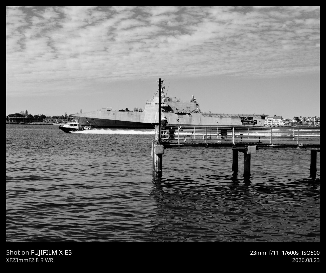 A man fishes from a dock in Coronado Bay, San Diego, while a massive naval vessel passes by.