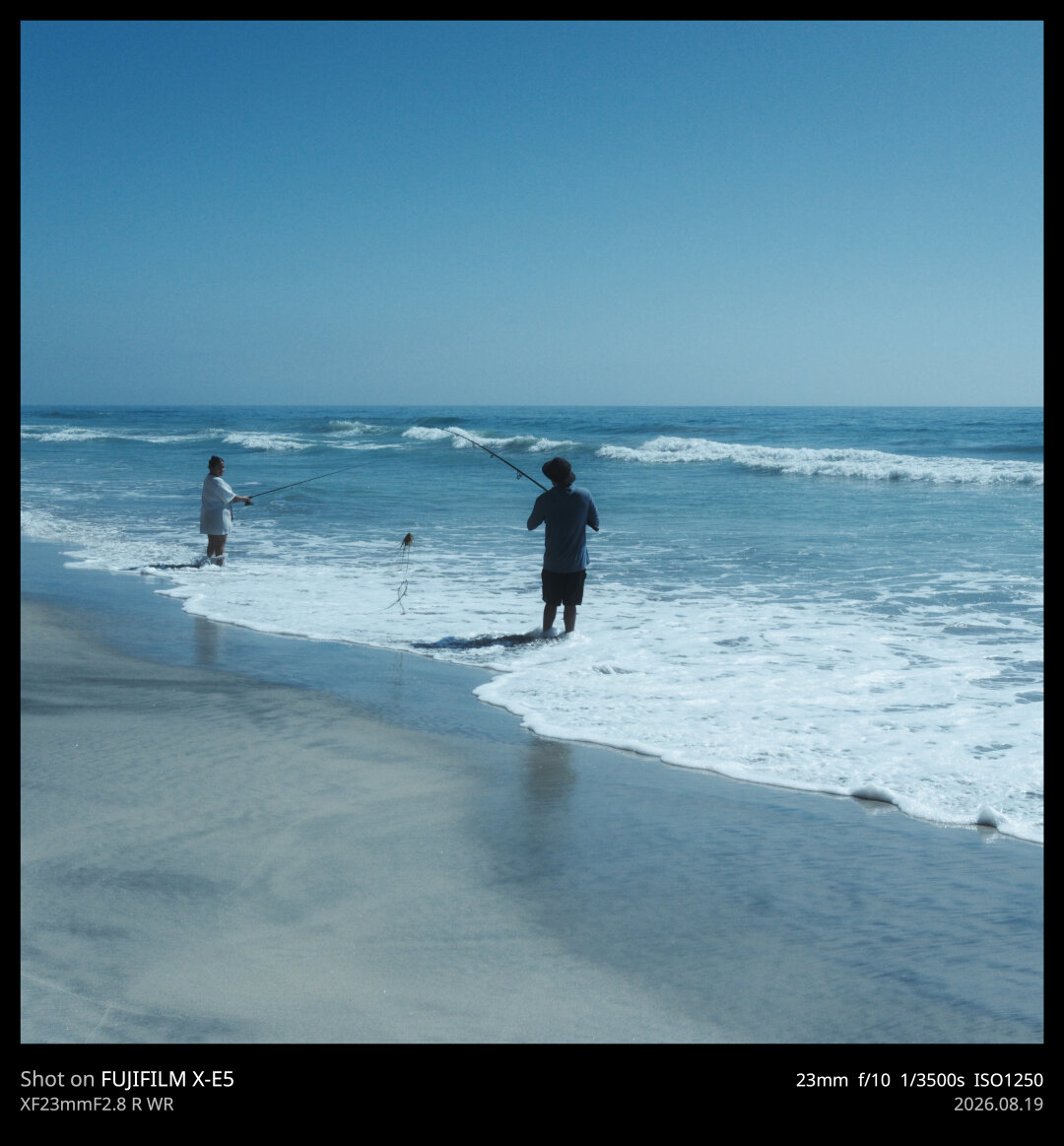 A man and his wife (presumably) are long line fishing while standing on the coast. The man has just reeled in his catch: a cluster of seaweed.
