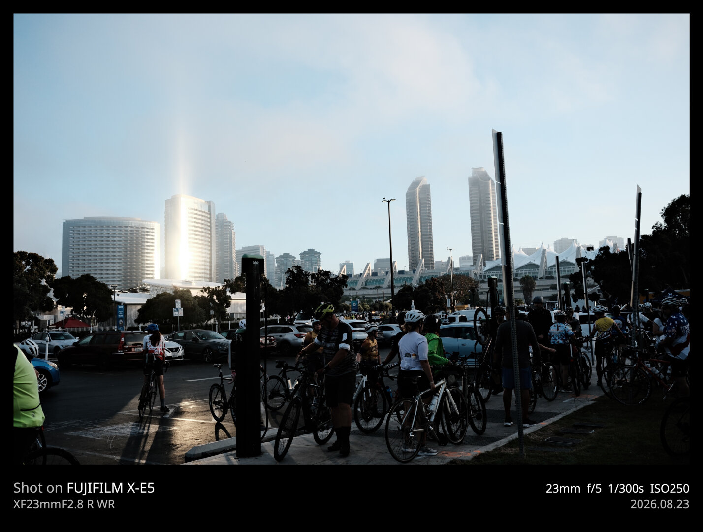A large group of bicyclists prepare to start a ride in the foreground. In the background, a tall building reflects the sunrise, creating a brilliant spire of light, which also illuminates part of the parking lot where the bicyclists are gathered.