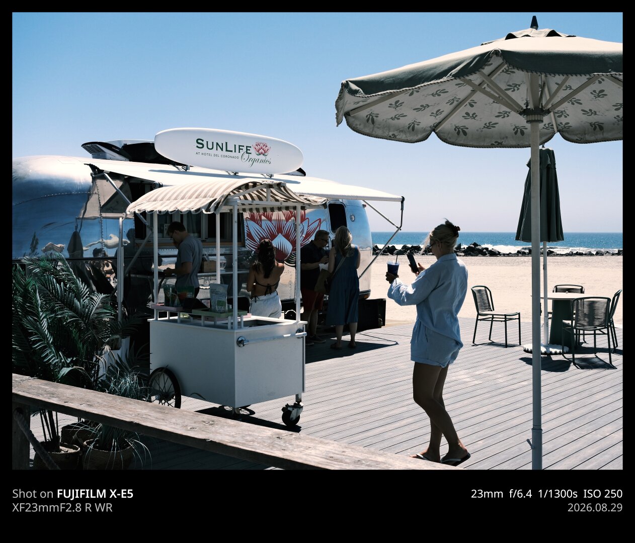 A woman is taking an iPhone photo of her smoothie, which she has purchased from a beach side establishment called "SunLife." The establishment is what appears to be an Air Stream trailer, with a very shiny metal surface. In the background you can see the Pacific Ocean and Coronado Central Beach.