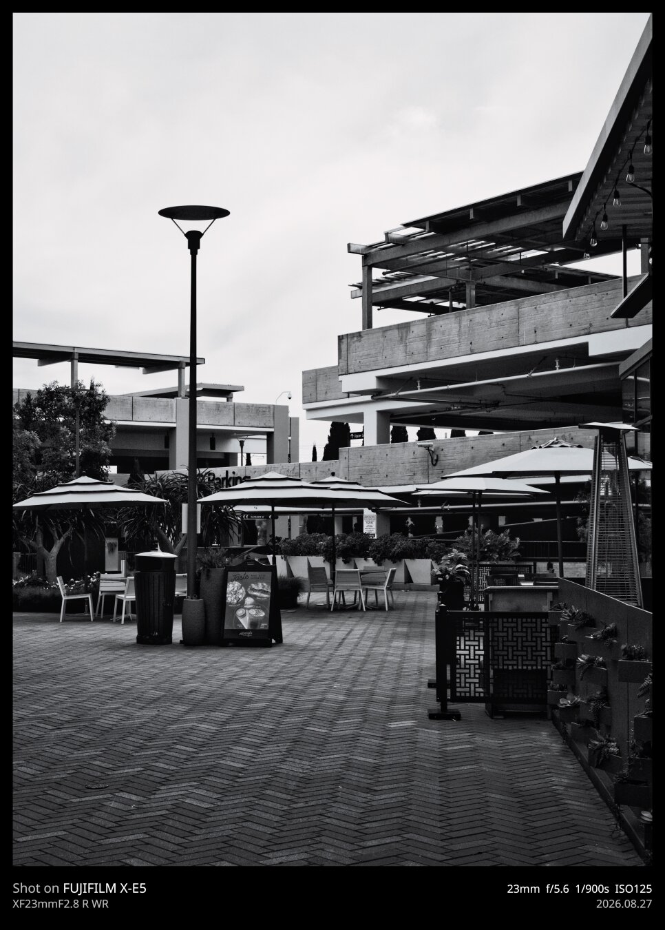 An empty plaza in an outdoor shopping mall. There are many sun umbrellas hiding people who are not there from sun that is not present because it's overcast. The scene is dominated by parking structures and herringbone walkways.