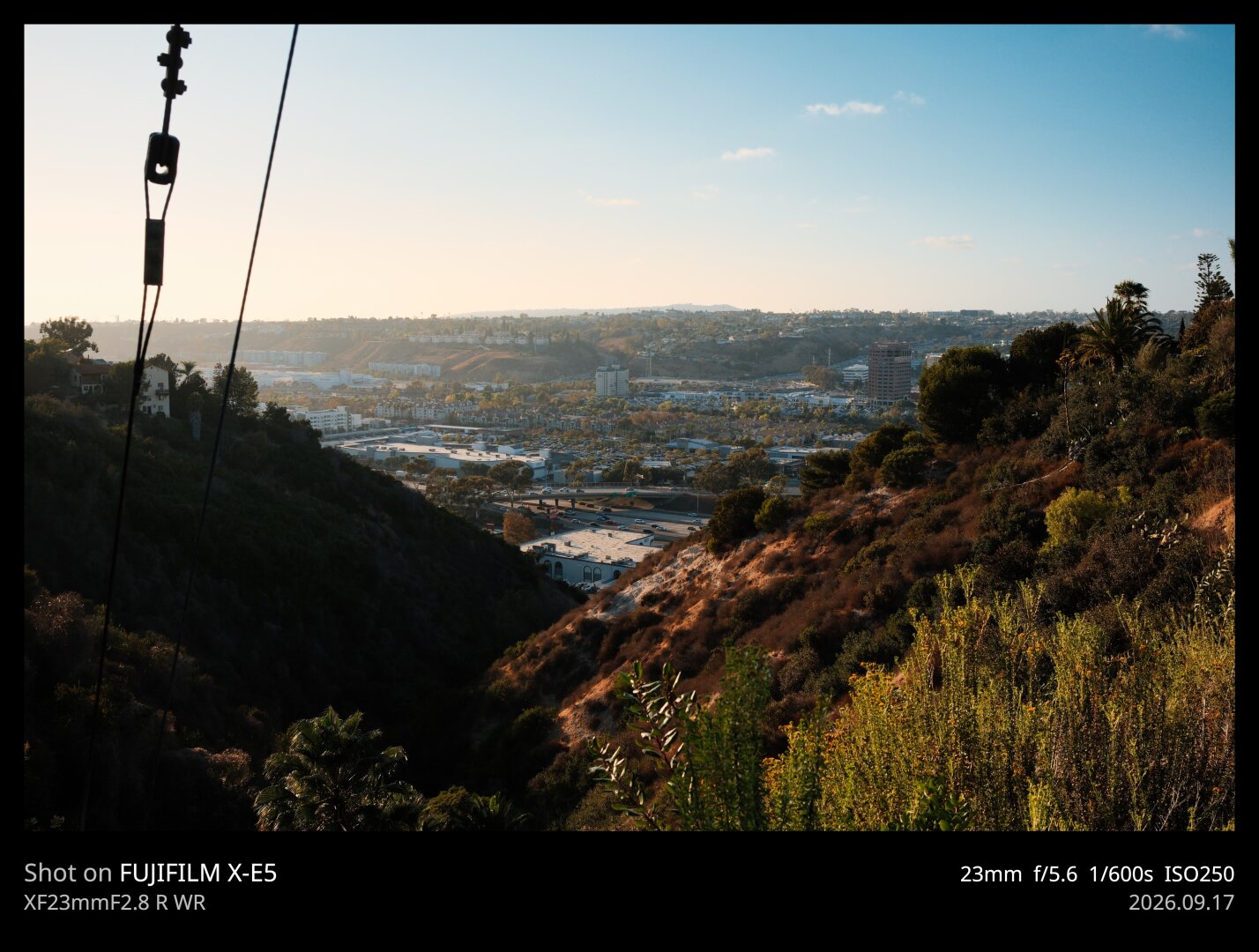 View from a the end of a dead end rode in University Heights. You see a canyon with southwestern style brush, emptying out into a highway and the roofs of shopping plazas in the middle distance. The sun sets off camera left, throwing soft golden light onto the scene.