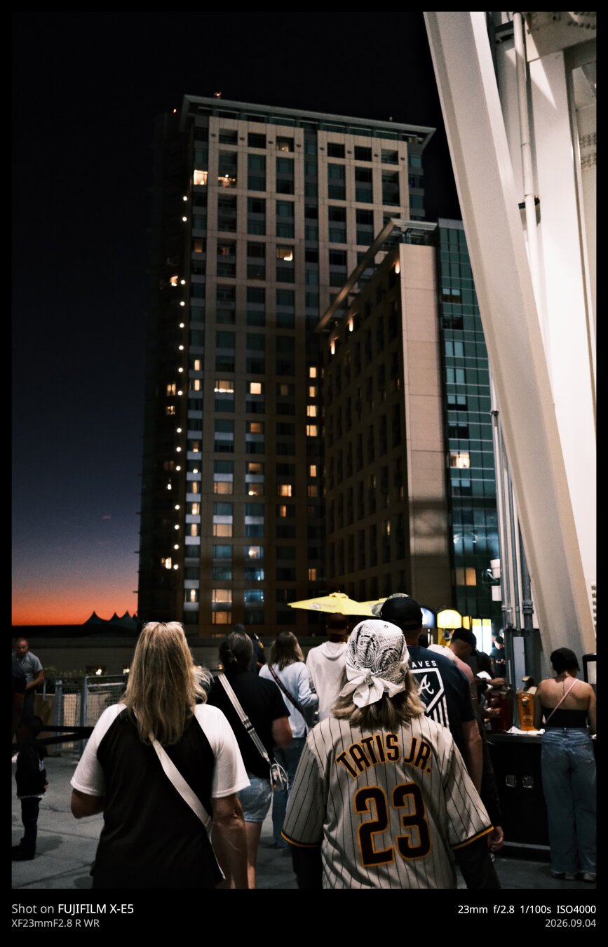 photo taken from a walkway in Petco Park, home of the San Diego Padres. Two women are in the foreground, their backs to the camera. One is wearing a Tatis Jr jersey. The sun is fully set in the background, with just a bit of orange and purple illuminating the sky. An apartment building is in frame, partially lit by the stadium lights.