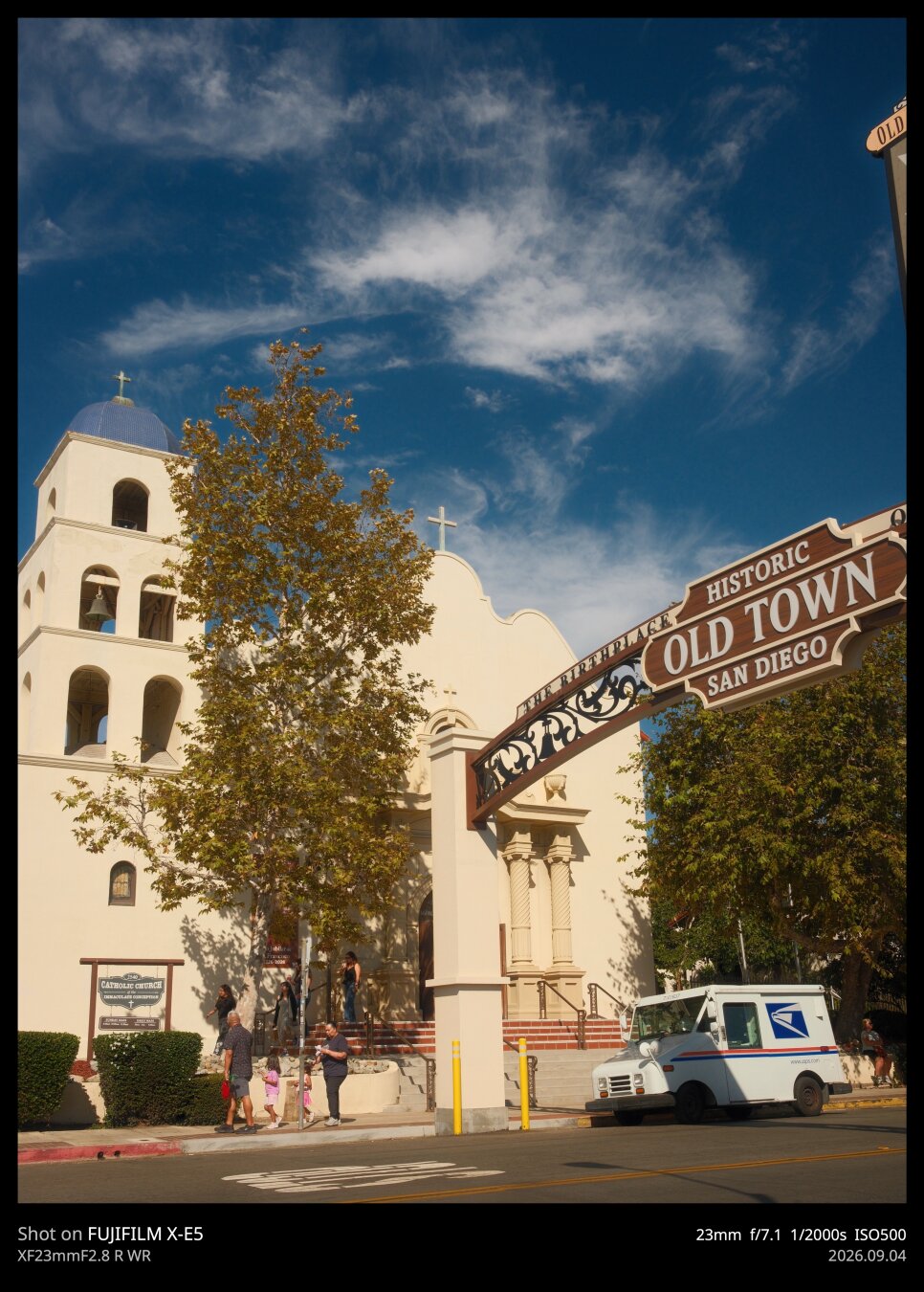 A photo of the Old Town neighborhood archway, which sits almost directly in front of the old Immaculate Conception Spanish style Catholic Church. The sky is a deep blue with a few wispy clouds. Outside of the church, a postal van is parked.