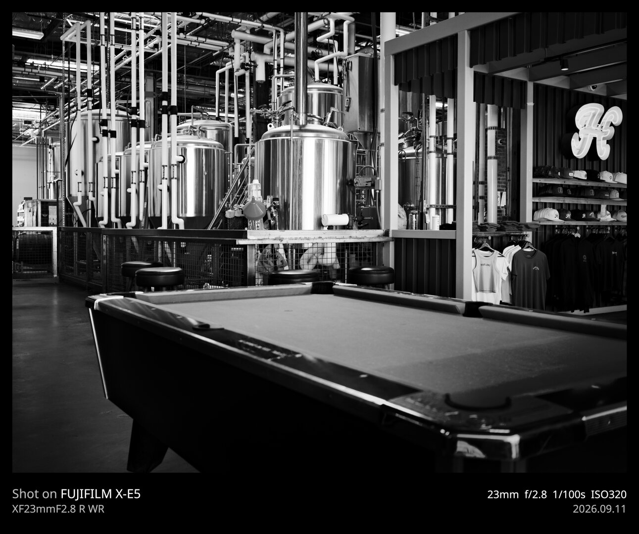 A high contrast black and white photo of the inside of a microbrewery. In focus in the background stand the large, shiny metal cylinders the brewing takes place in. In the foreground, and slightly out of focus, is a pool table. On the right side of the frame is a large Harlan logo (a stylized 'H') in the gift shop.