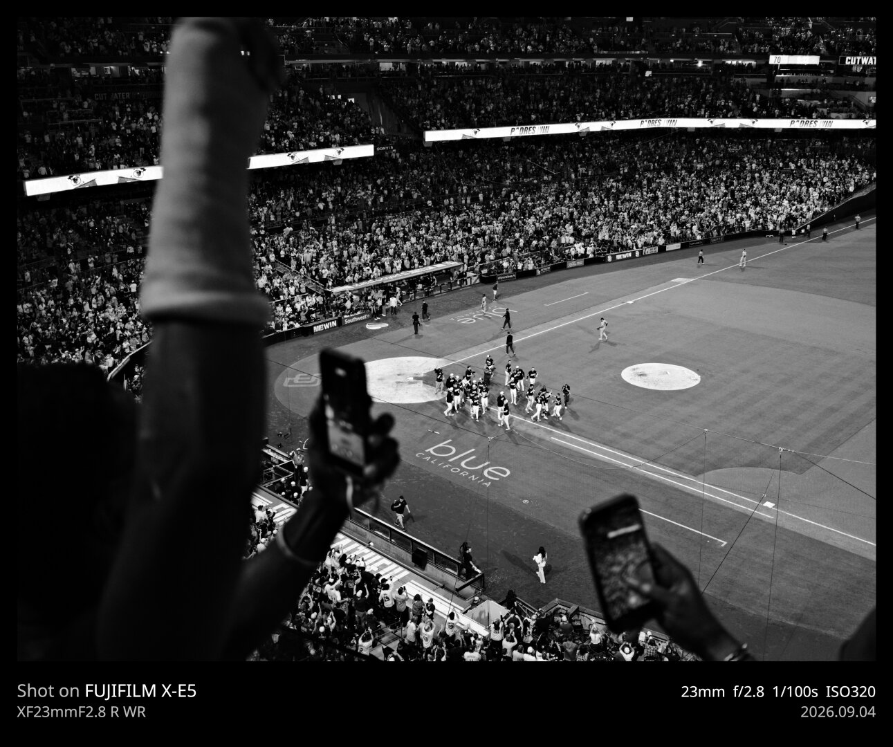 Black and white photo of the infield of Petco Park, taken from the upper deck, 1st base side. The players are celebrating their win around home plate. In the foreground, fists are pumping and people have their phones out recording the moment.