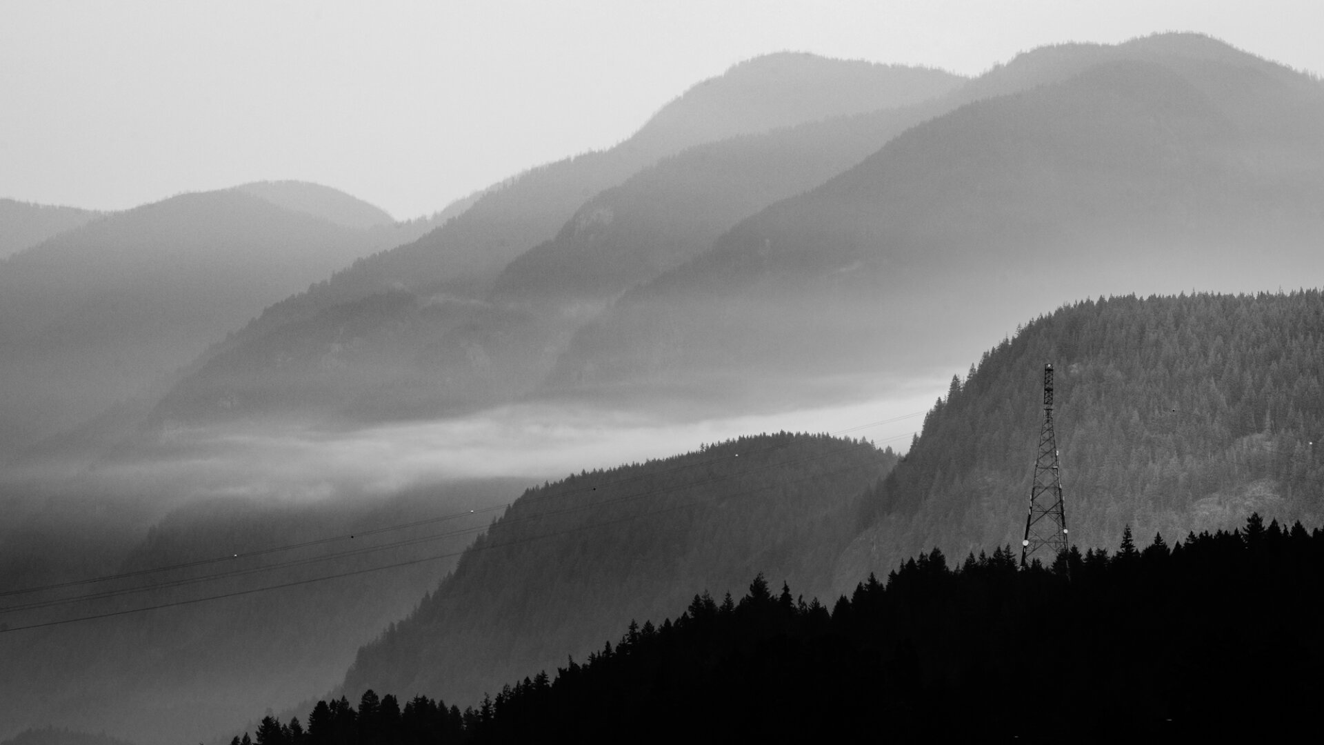 A black and white photo of the mountains above Belcarra, BC. It is a smoky morning, which lends a somber mood to the shot. A tower connecting powerlines from shore to shore is visible, with a thin, dense line of clouds stretching out from behind it's hill. A dark line of forested mountain fills the foreground.
