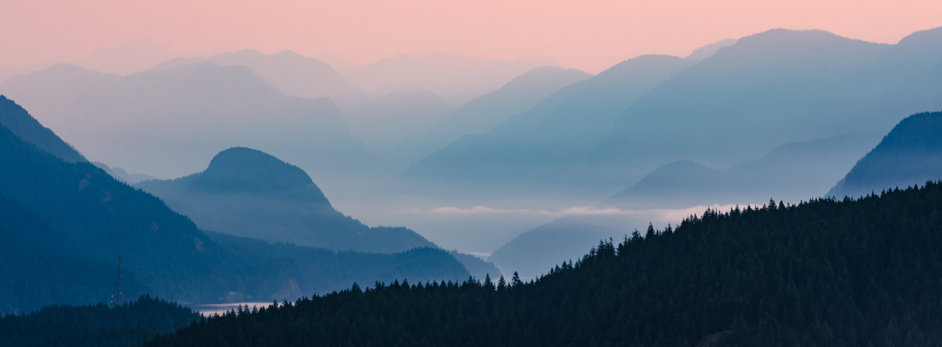 A landscape photograph of the mountains north of Burnaby Mountain, in Vancouver, BC. It is sunrise on a morning filled with wildfire smoke, which gives an ominous purple and blue tint to the scene. The smoke highlights the layers of mountains going up Indian Arm, with a familiar powerline tower visible in the foreground.
