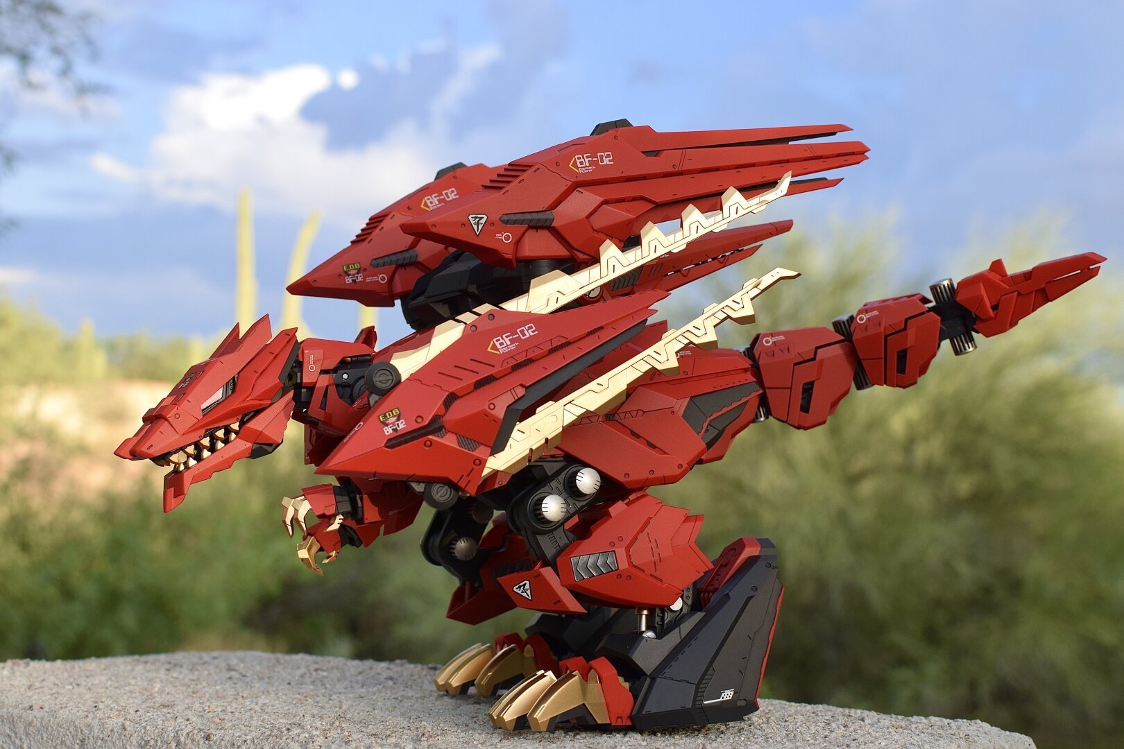 A detailed red robotic t-rex figure stands on a surface, showcasing metallic features and intricate design elements. The background features a natural landscape with soft clouds and green vegetation.