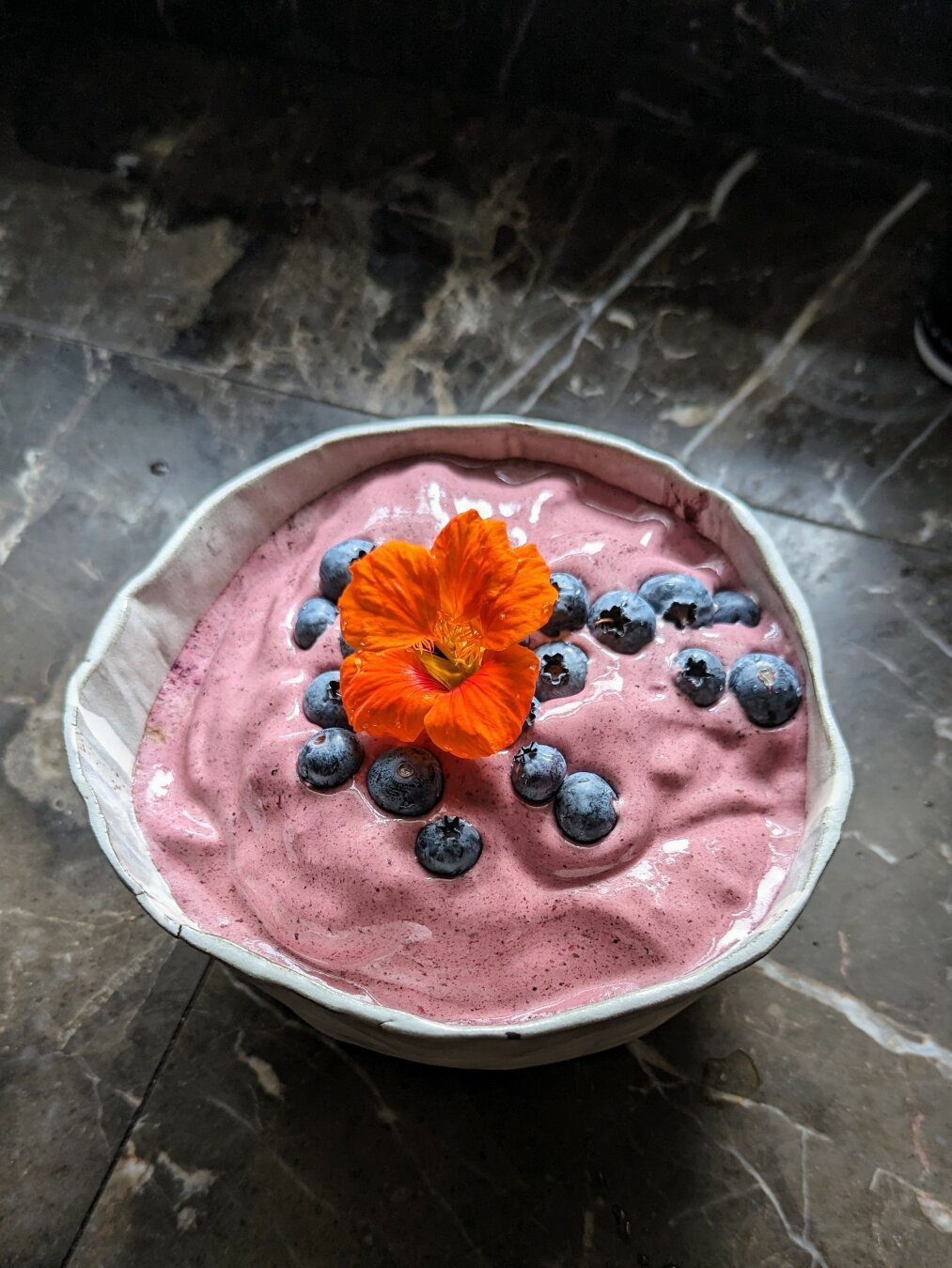 A light grey bowl on a marble background. The bowl contains a purple smoothie mixture topped with fresh blueberries and an orange nasturtium flower.