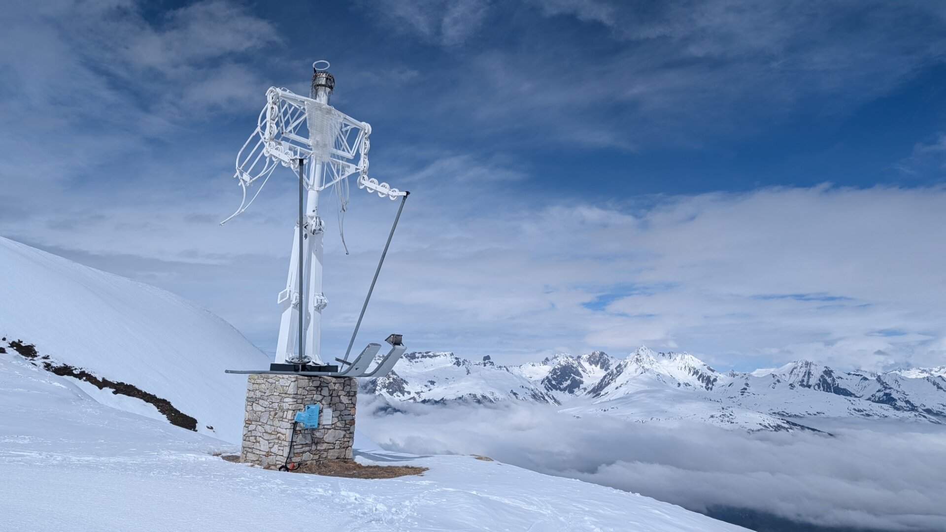 A skiing angel made of old ski lift parts stands on a mountain top, there are other mountains in the distance above a sea of clouds