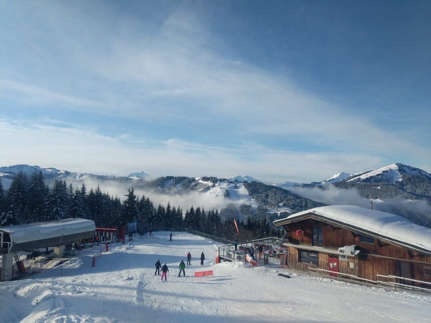 snowy ski slopes, a building and the bottom station of a ski lift surrounded by trees. there are snowy mountains in the distance and light clouds in a blue sky