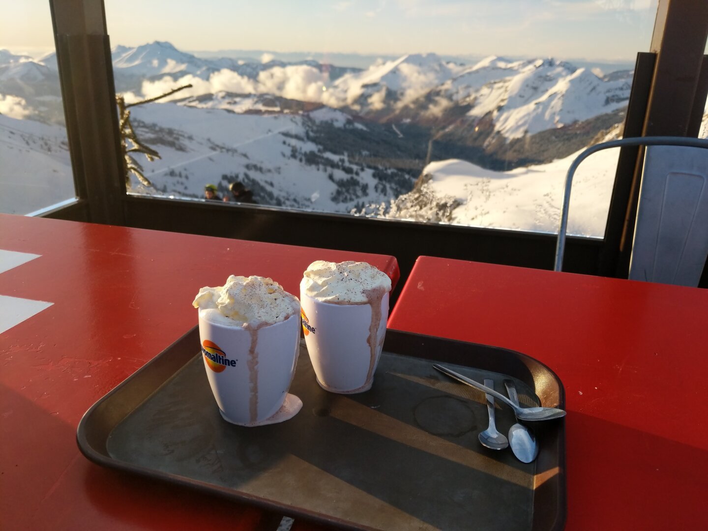 2 hot chocolates on a tray on a table. behind the table the window has a view down from the top of a snowy mountain into a valley