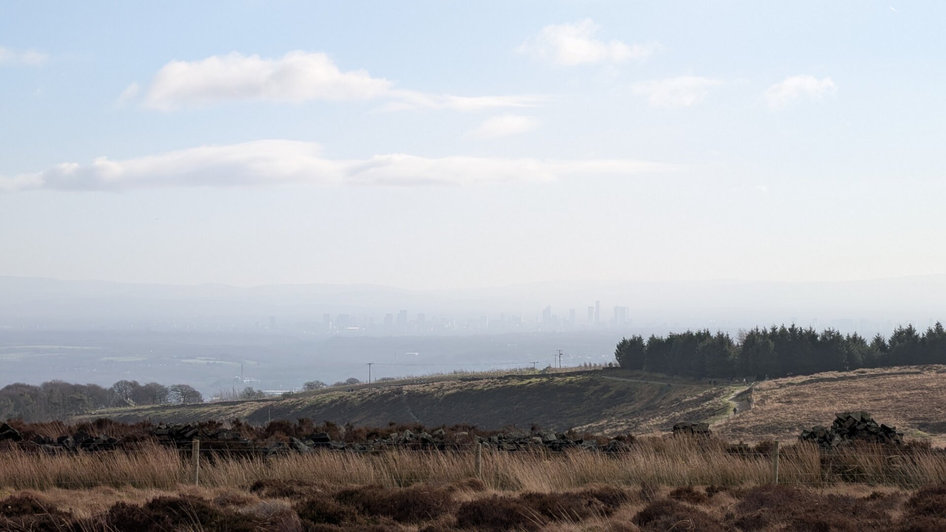 A hazy view of Manchester from a hillside about 20 miles away. The sky is blueish with some small clouds