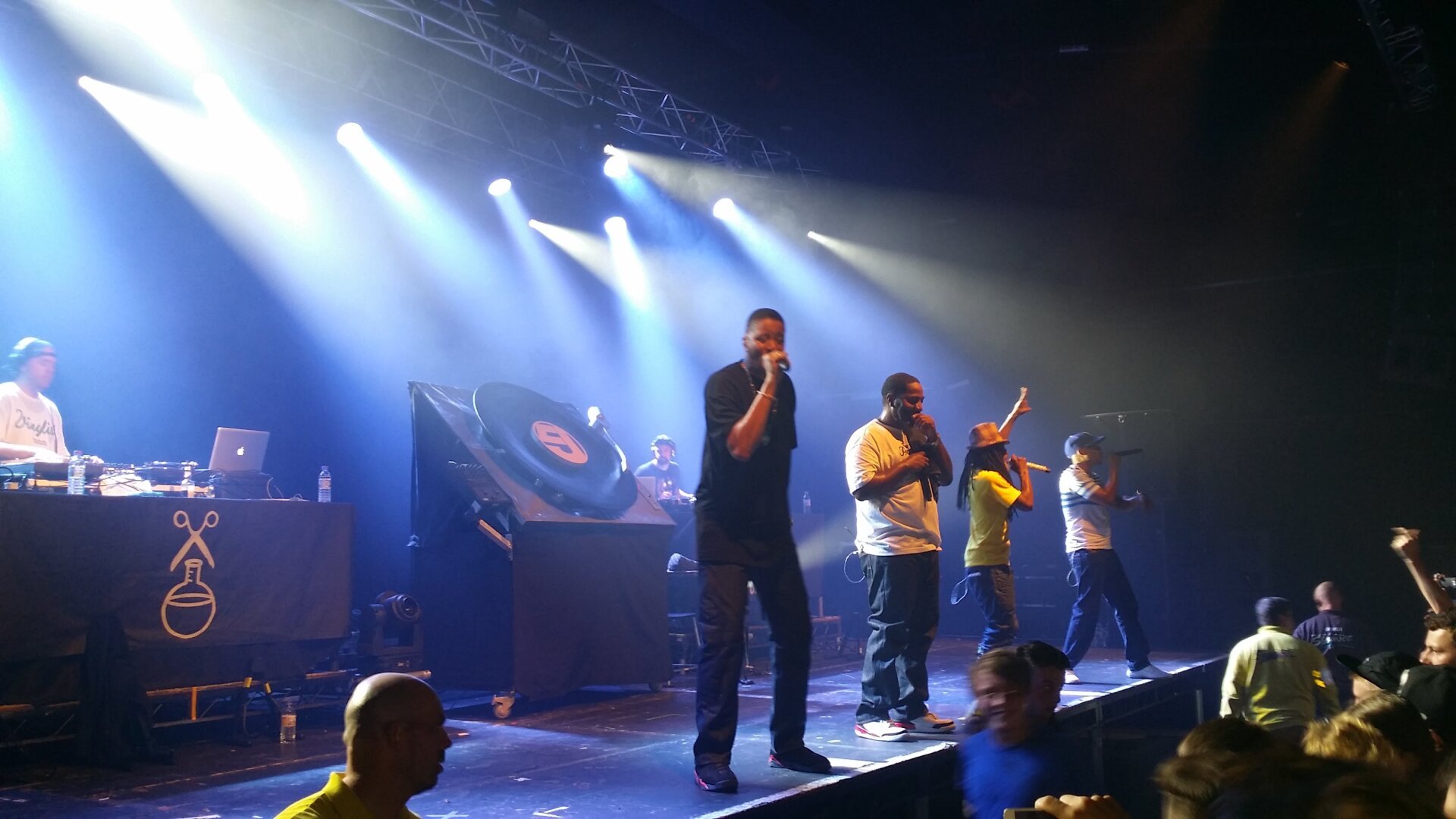 A group of four rappers on stage with two DJs, with lights shining out into the crowd