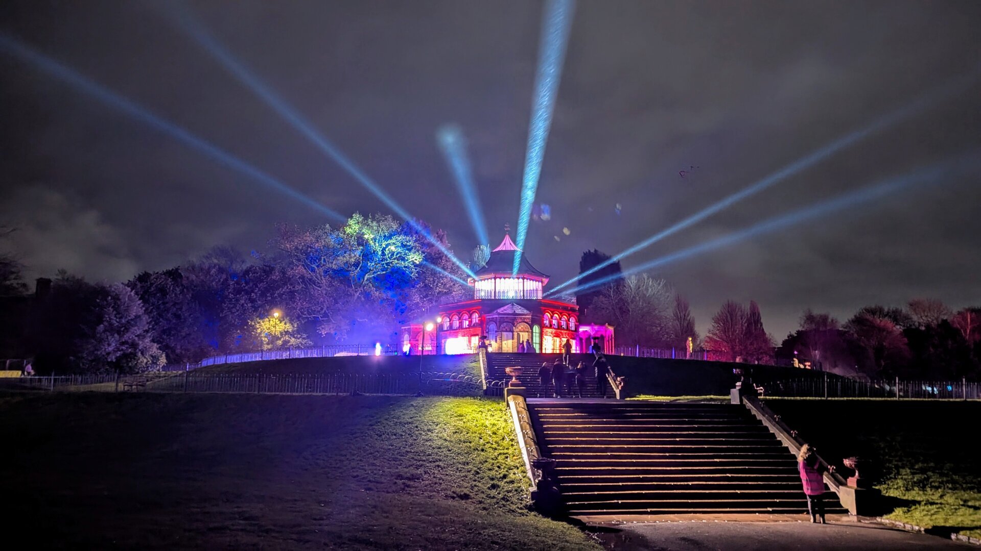 A building at the top of a small hill with steps leading up to it. spotlights are coming out of the glass dome at the top of the building. it is night time.