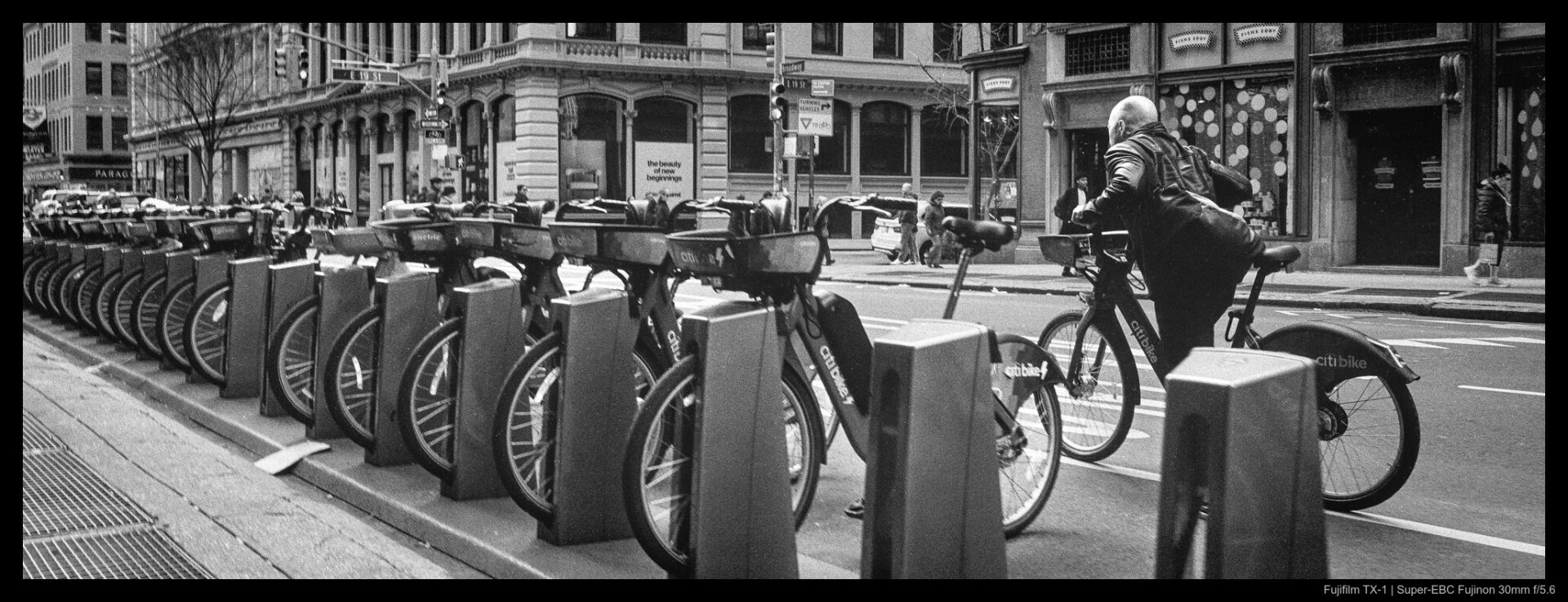 Someone renting a Citibike prepares to ride, mounting up while facing down a line of other docked bikes parked on the side of a city street.