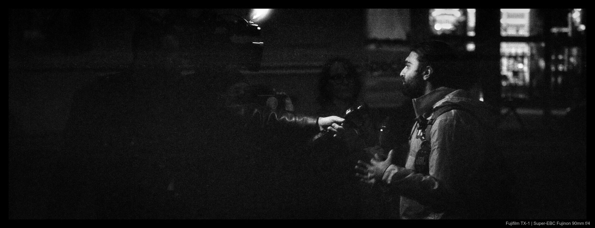 A protester is interviewed by a cameraman shrouded in darkness, lit by an LED panel from above. The interviewer's hand is visible emerging from the shadows, holding a microphone towards the speaker.