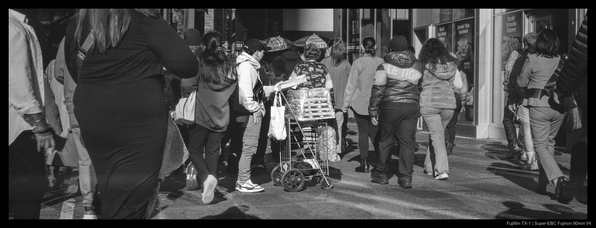 A street vendor with a stack of churros on a push cart stands in the middle of a crowd that parts around them a second glance.