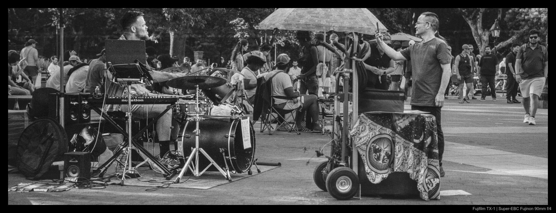 A person holds their phone up to record a drummer sitting in the middle of their equipment, their full kit set up tucked against one of the curved stone benches in Washington Square Park.
