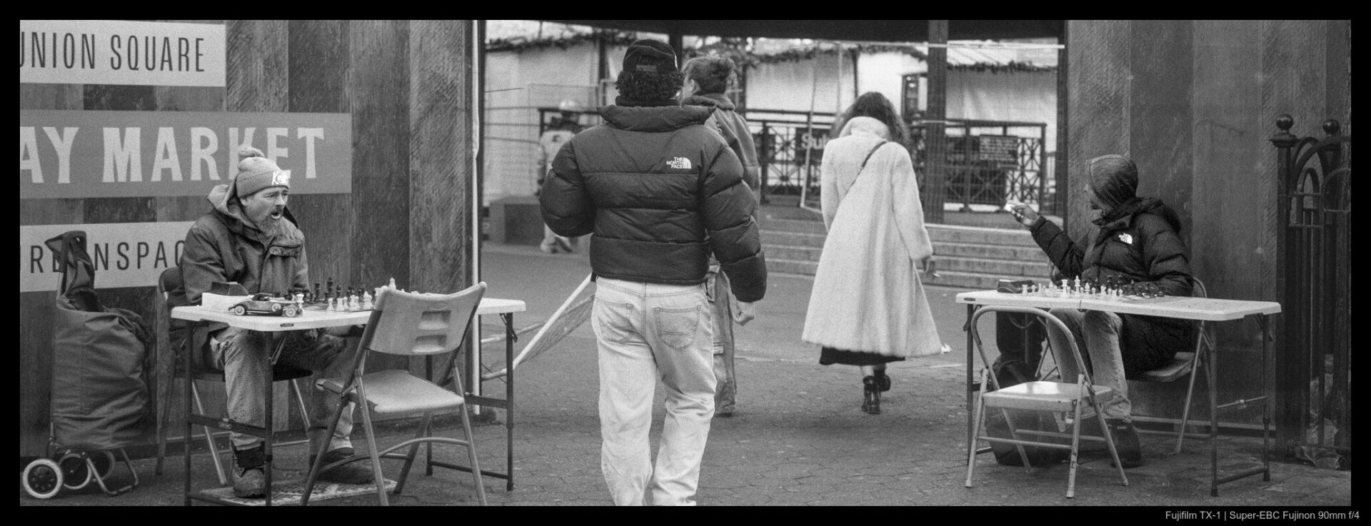 A trio of pedestrians pass through stalls in the Union Square Holiday Market, flanked on both sides by chess players seated at folding tables.