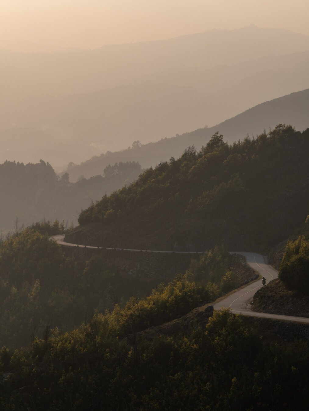 A photograph of a landscape, with a twisty road carved through the hills, in a foggy afternoon. A motorcycle can be seen on the road.