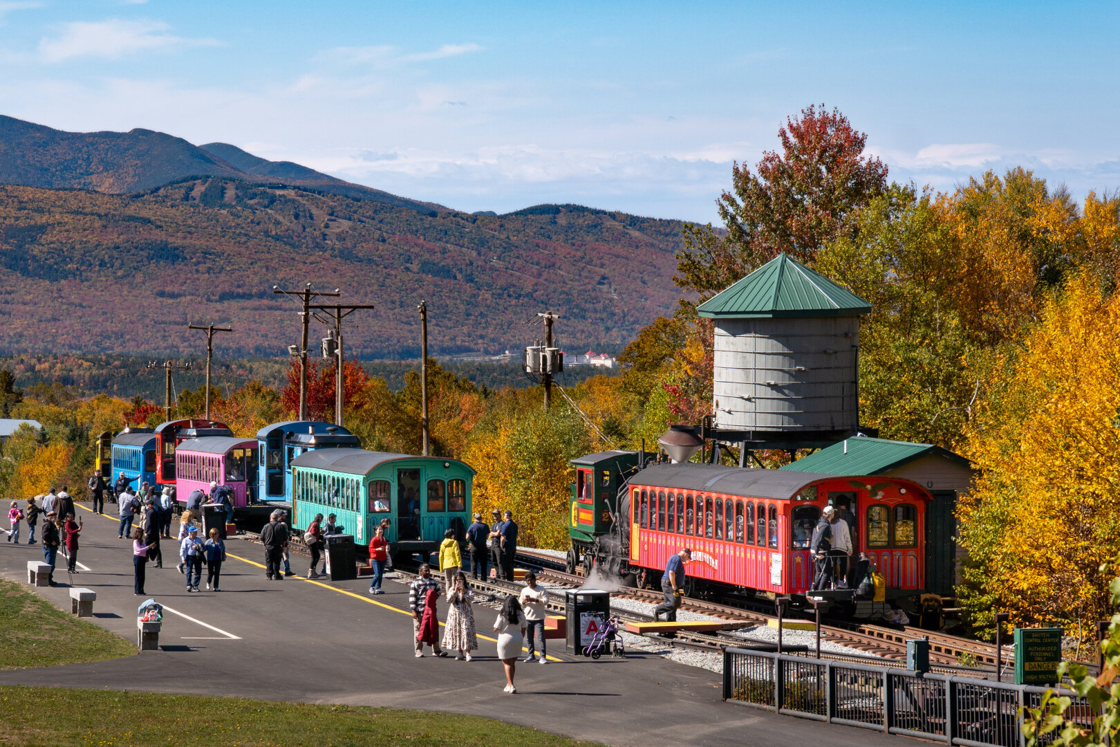 A rural train station with train cars and locomotives in the foreground.
Mountains with fall colors in the background.