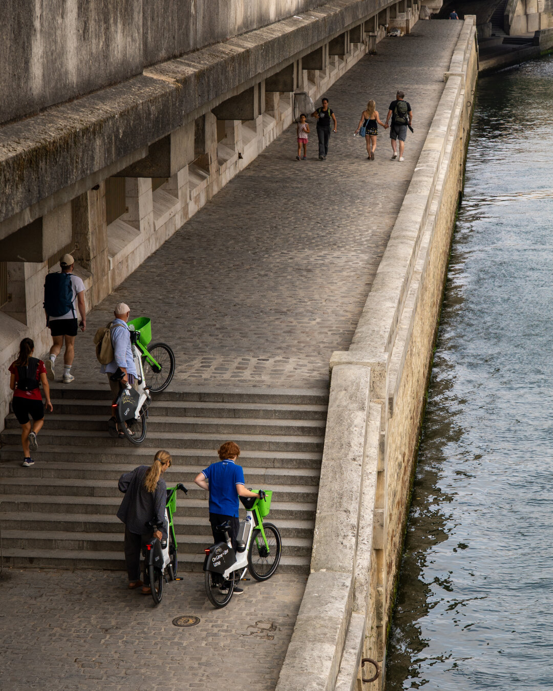 People walking by the Seine river in central Paris. Some are hauling their bikes over a set of stairs.
