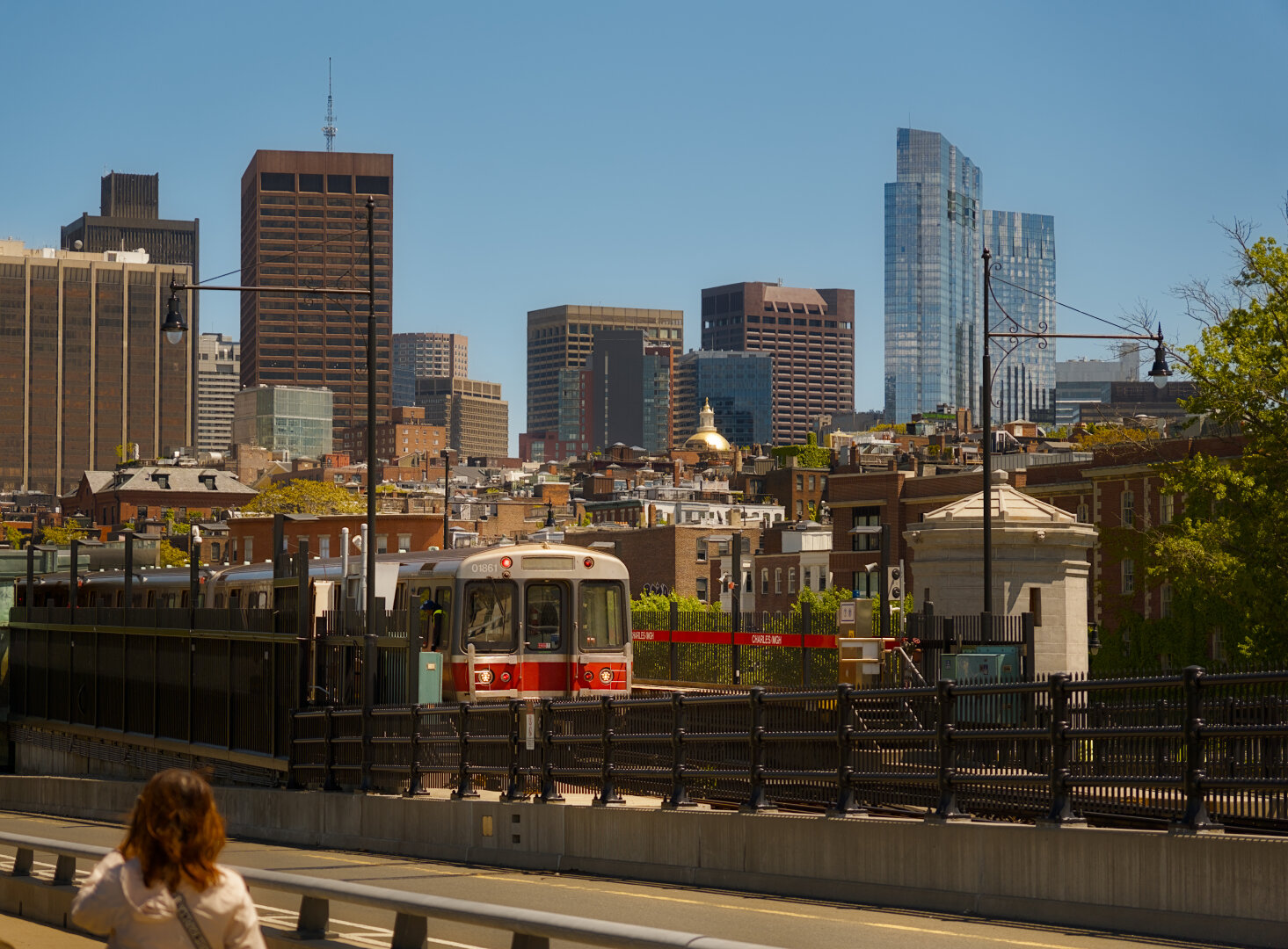 In the foreground, a red-line train is stopped at a station. In the background, downtown-Boston's skyline.