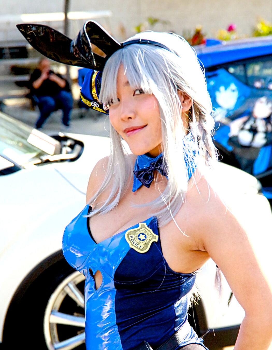 Young asian girl in a blue bunny outfit cosplaying as Judy Hopps from Zootopia.  Black bunny ears, hair covers one eye, lots of cleavage and bare shoulders and armpit.  Blue bunny outfit has the police badge and belt.