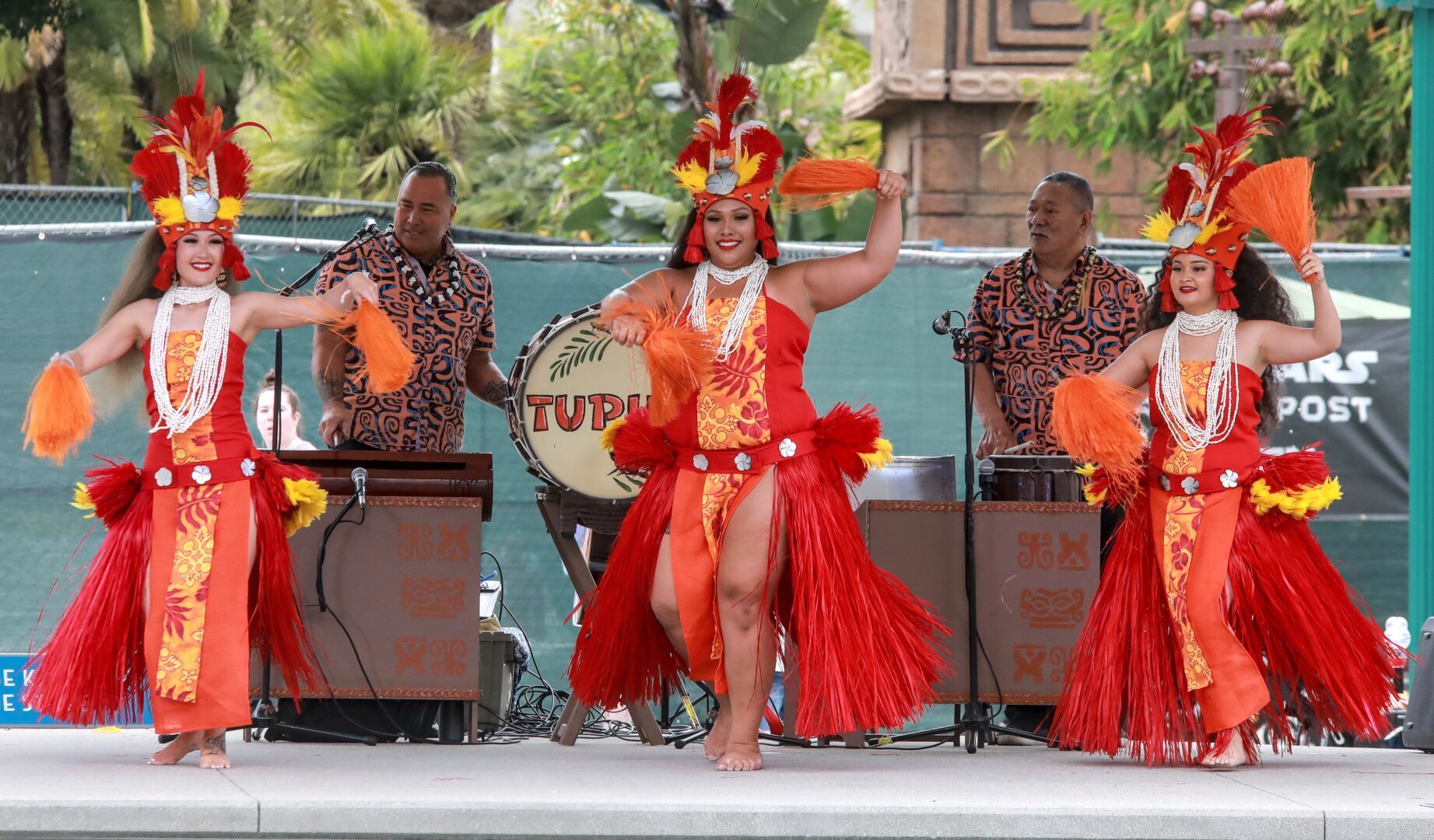 Three Tupua polynesian dancers in their orange and yellow attire.  Hips are swaying and the legs of the middle performer are visible through the grass skirt.  The have their arms up ans they perform.