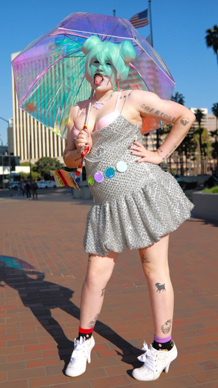 A young person in costume as Bubble from the Amazing Digital Circus.  She is holding an umbrella which gives a soap bubble coloration.  Her teeth have sharp points and the tongue is out with a metal piercing which provide a menacing look.  The top is very low cut and there is plenty of cleavage visible.  Her hand is close to face and the fingers are in a threatening position.