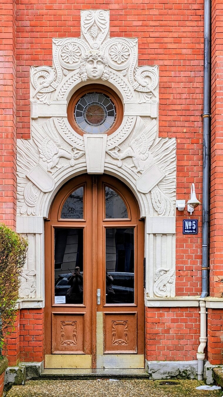 A set of arched wooden doors on a red brick building, topped by an incredibly ornate white stone carving featuring a central face, circular windows, and two phoenixes.