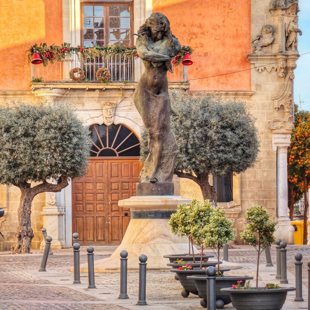 A warm, late afternoon view of a bronze statue of a woman with flowing hair standing on a stone pedestal in a cobblestone plaza. Behind the statue is a large wooden double door with a radiating fanlight above it, set into a building with an orange-pink facade. Above the door, a balcony adorned with festive red bells and green garlands hangs below a window. To the left and right of the statue are mature olive trees with rounded canopies. In the foreground, several dark gray planters with small green plants and a few pink flowers are arranged around the base of the statue, interspersed with short, dark bollards. On the far right, a tree laden with bright orange fruit is visible. The sky is clear blue. A warm, late afternoon view of a bronze statue of a woman with flowing hair standing on a stone pedestal in a cobblestone plaza. Behind the statue is a large wooden double door with a radiating fanlight above it, set into a building with an orange-pink facade. Above the door, a balcony adorned with festive red bells and green garlands hangs below a window. To the left and right of the statue are mature olive trees with rounded canopies. In the foreground, several dark gray planters with small green plants and a few pink flowers are arranged around the base of the statue, interspersed with short, dark bollards. On the far right, a tree laden with bright orange fruit is visible. The sky is clear blue.
