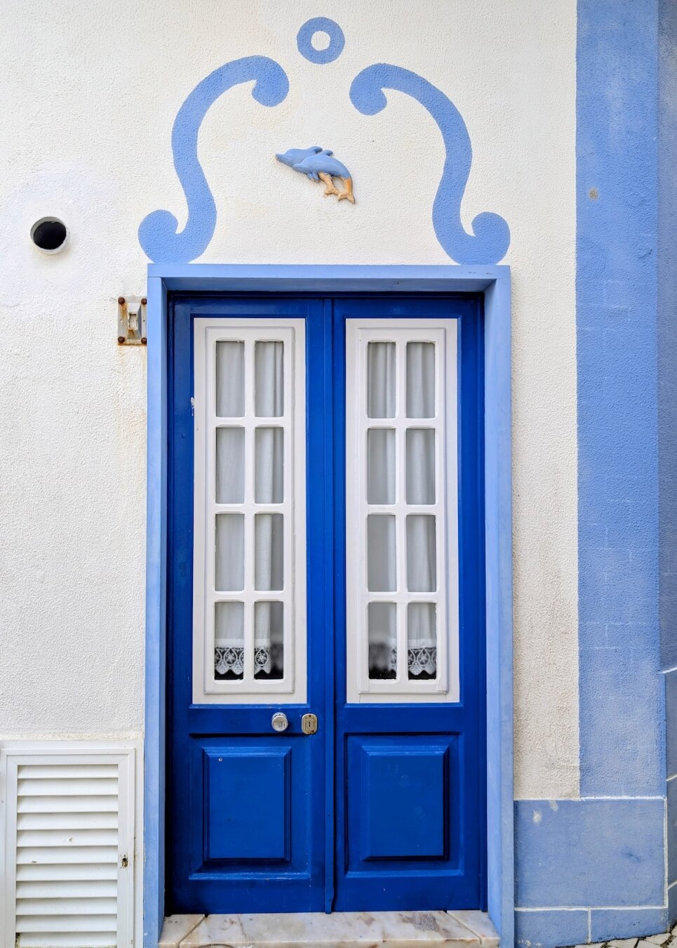 Vibrant blue door with two white-framed glass panels, featuring a blue painted scroll design and a small dolphin figurine on the white wall above the frame.