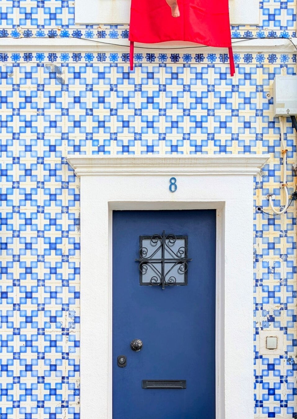A solid navy blue door with a small decorative iron grate and a black knocker, set against a wall covered in traditional blue and white geometric Portuguese tiles.