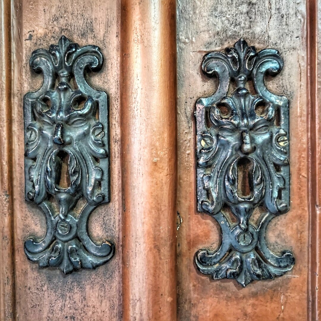 A close-up of two identical dark metal keyhole covers on a wooden door, each intricately cast in the shape of a bearded, mythological face with flowing hair.