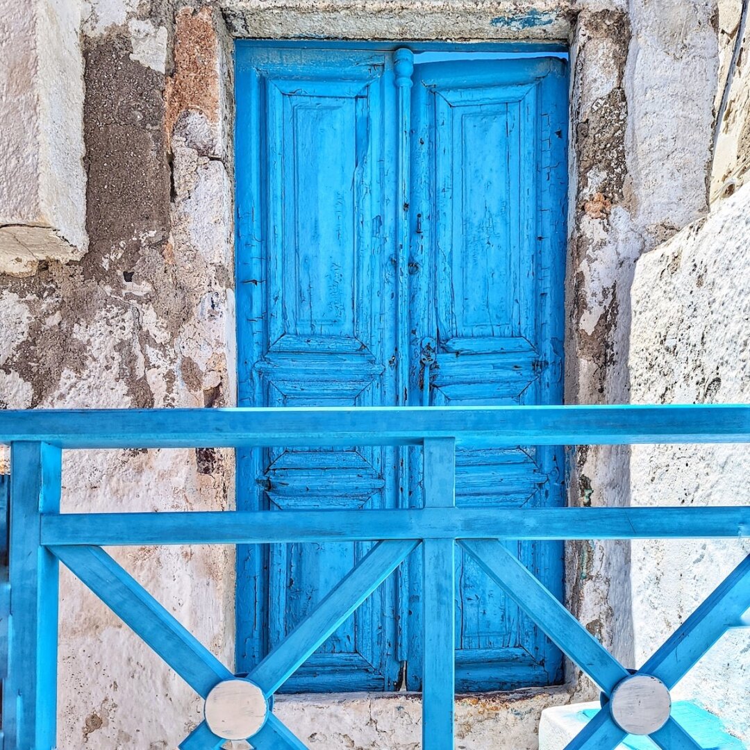 Weathered sky-blue wooden double door with peeling paint, set into a whitewashed stone wall behind a matching blue crossbar railing in Santorini, Greece.