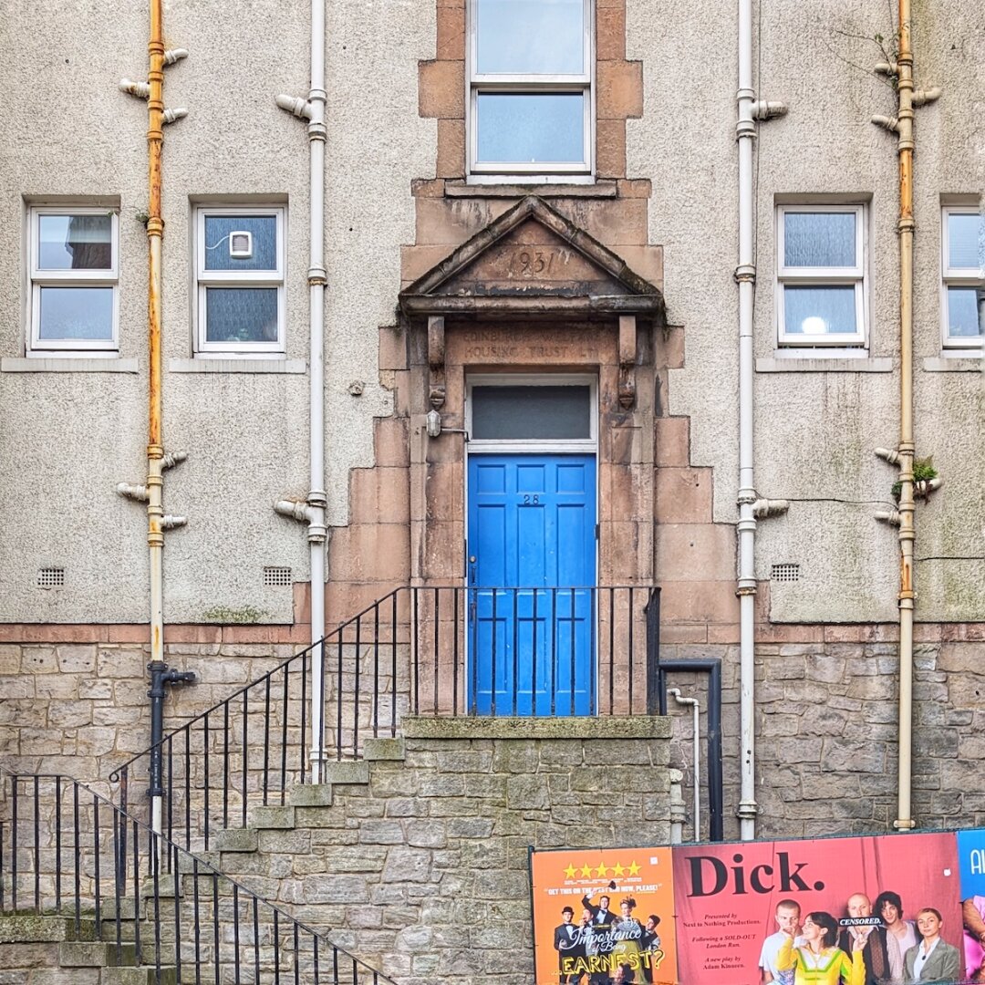 A bright blue entrance door at the top of stone steps, set beneath a 1931 carved stone pediment with Edinburgh Fringe show posters displayed at street level.